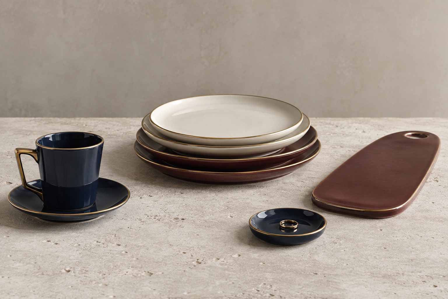 A collection of navy cups and saucers, ivory and burgundy plates, a ceramic serving board, and a small jewelry dish arranged on limestone.